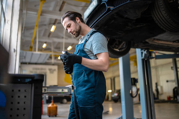 Serious focused young Caucasian automobile mechanic looking at the drill and nozzle in his hands