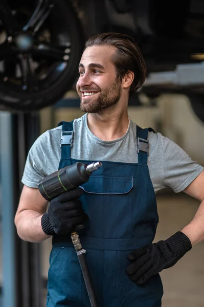 Glimlachende Jonge Auto Reparateur Houdt Een Boor Hand Kijkt Weg — Stockfoto