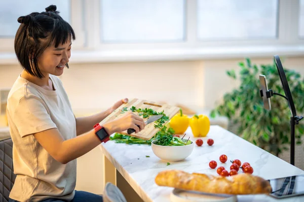 Joyous vlogger seated at the kitchen table adding the chopped parsley to the ceramic salad bowl while recording her vlog