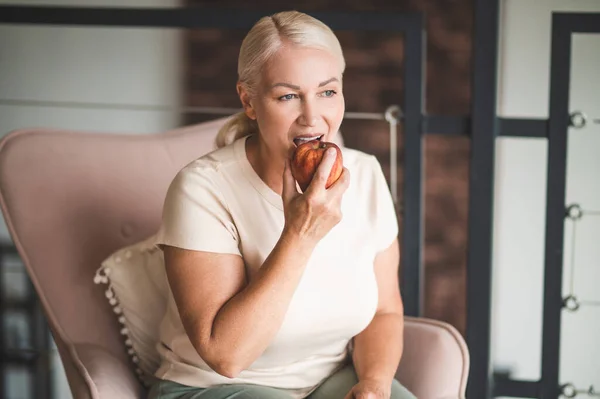 夢のような魅力的な成熟したブロンド女性座っていますでザレザーArmchair食べるAアップルで彼女の手 — ストック写真