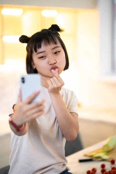 Young female making a finger heart gesture in front of the smartphone camera while taking a selfie