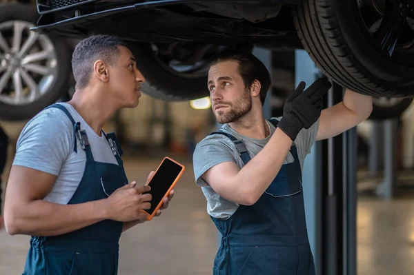 Automotive technician pointing at the automobile wheel to his colleague during the routine car inspection