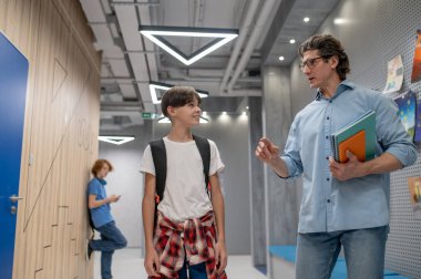 Okul arkadaşı bir öğretmenle konuşurken akıllı telefonlu bir çocuk koridorda duvarın önünde dikiliyor.