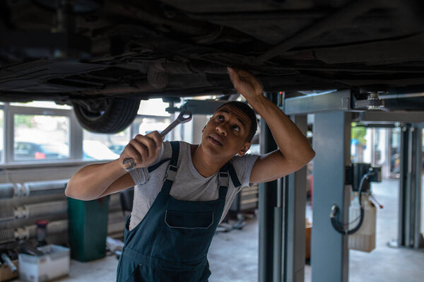 Focused mechanic holding a wrench in the hand and looking up at the vehicle underside