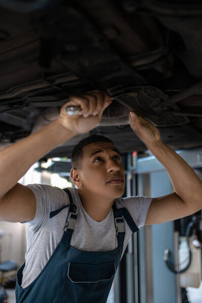 Concentrated African American car technician tightening the nuts on the automobile underside with a wrench