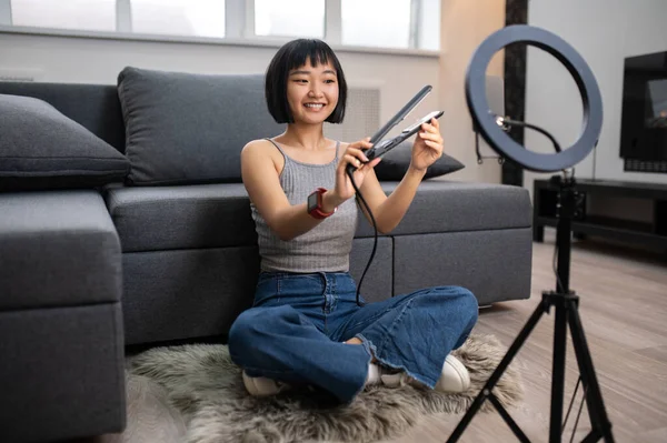 Smiling vlogger seated cross-legged on the floor rug holding a curler before the smartphone camera