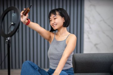Akıllı telefon kamerasının önünde oturan memnun vlogger elinde kozmetik yağı şişesi tutuyor.