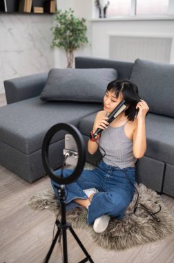 Vlogger halıya oturmuş, akıllı telefon kamerasının önünde saçını bigudiyle şekillendiriyor.