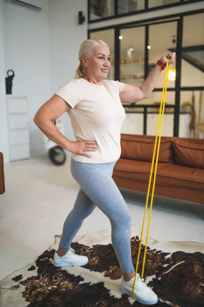 Smiling happy strong mature plus-size woman in sportswear performing the resistance band single-arm bicep curl