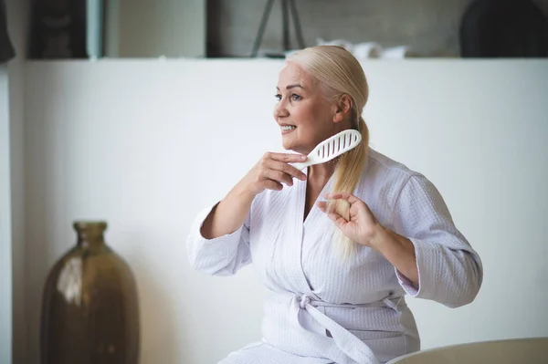 Smiling happy beautiful mature lady in the terry bathrobe combing her ponytail with the hairbrush