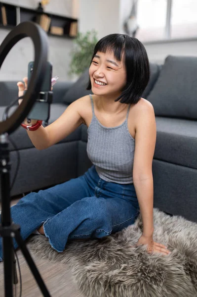 Cheerful Asian video blogger sitting on the floor rug while adjusting the settings on her smartphone
