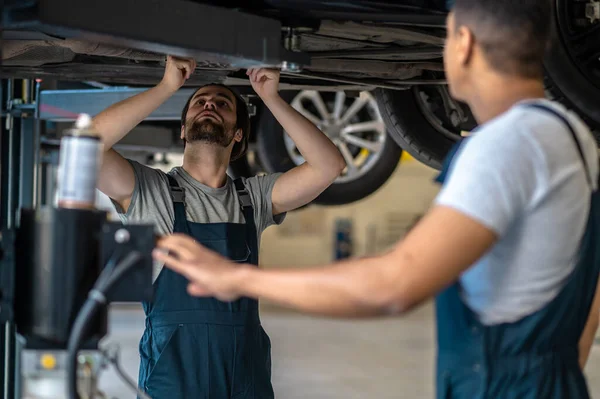 Focused experienced automotive technician performing visual inspection of the car underside assisted by his colleague