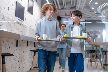 Neşeli öğrenci ve neşeli sınıf arkadaşı okul kafeteryasında yemek tepsilerini birlikte taşıyorlar.