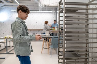 Okul kantininde. Okul çocuğu öğle yemeğinden sonra tepsisini mutfak raflarına koyuyor.