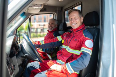 Gülümseyen neşeli bir sürücü ve kırmızı üniformalı bir erkek doktor ambulans arabasında oturuyor.