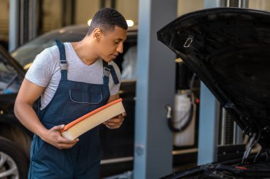 Bir otomotiv filtresi tutan Afrikalı Amerikalı tamirci açık araç kaputunun altına bakıyor.