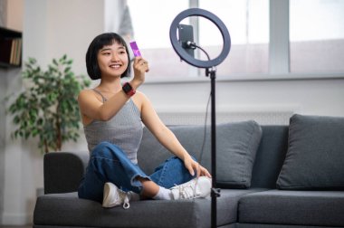 Neşeli vlogger koltukta bağdaş kurup akıllı telefon kamerasından önce elinde bir tüp kremayla oturuyor.