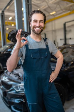 Gülümseyen neşeli oto tamircisi iş kıyafetleri içinde benzin istasyonunda duruyor ve 