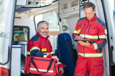 Ambulans arabasında oturan neşeli meslektaşının önünde tableti kullanan odaklanmış sağlık görevlisi.