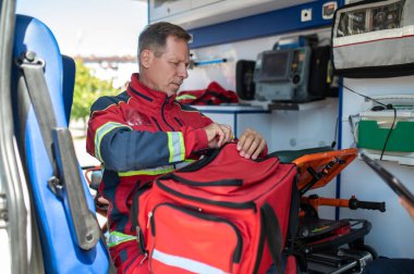 Acil yardım çantasını açarken ambulans vagonunda oturan kırmızı üniformalı deneyimli sağlık görevlisi.