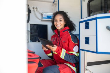 Elinde tablet olan neşeli bayan doktor ambulans vagonunda oturuyor.