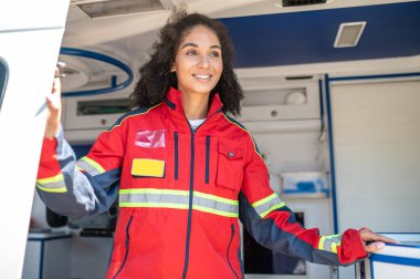 Gülümseyen bir kadın doktorun belden yukarı portresi ambulans arabasından uzaklığa bakıyor.