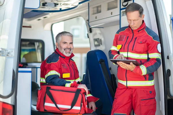 구급차에 즐거운 동료들이 자리에서 태블릿을 사용하는 집중적 요원들 스톡 사진
