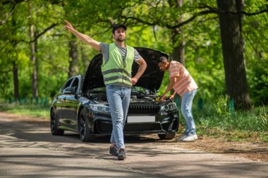 Yardım gerekiyor. Yeşil yelekli bir tamirci orman yolunda otostop çekiyor.