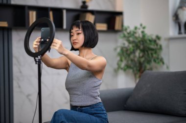 Odada oturan ciddi bir konsantre video blogcusu akıllı telefonu tripod standına koyuyor.