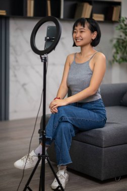 Akıllı telefon tripoda monte edilmeden önce kanepede oturan mutlu genç vlogger