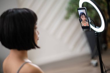 Akıllı telefonun önünde oturan düşünceli video blogcusu LED halka ışığıyla kürsüye monte edilmiş.