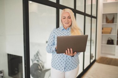 Gülümseyen stil sahibi olgun bir bayan duvarın yanında duruyor ve elindeki dizüstüne bakıyor.