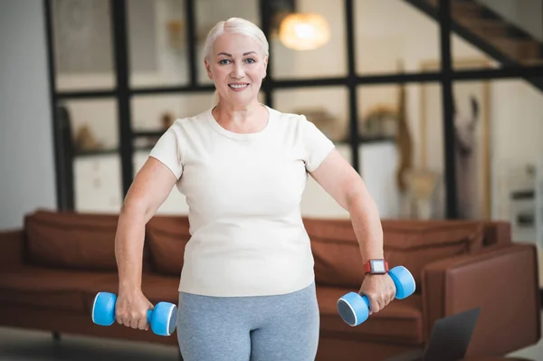 Smiling joyful attractive mature lady working out with a pair of hand weights at home