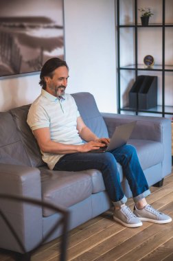 Evden çalışıyorum. Kanepede dizüstü bilgisayarla oturan yakışıklı olgun bir adam.