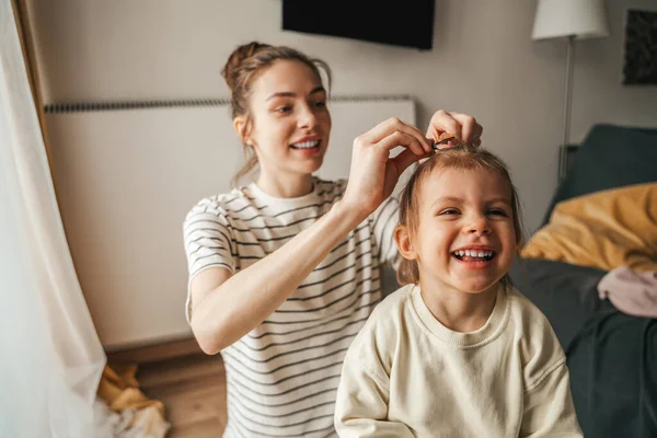 Smiling female parent securing hair of her pleased cute little child with a hair clip