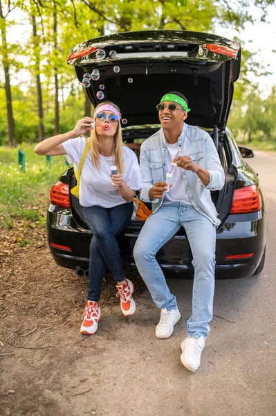 Bubbles. Cute couple sitting on the car boot and blowing bubbles