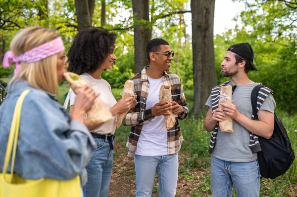 Lunch in the forest. Group of young people in the forest standing near the car and eating sandwiches