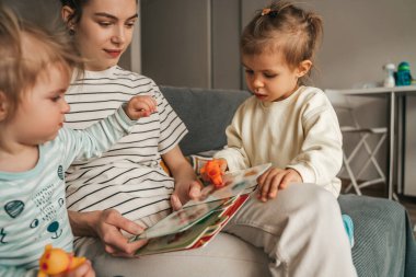 Çocukları oyuncaklarla oynarken elinde kitapla kanepede oturan hayalperest genç kadın.