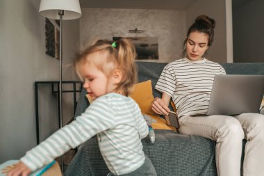 Dizüstü bilgisayarı olan bir kadın, çocuğu tek başına oynarken akıllı telefondan mesaj okuyor.
