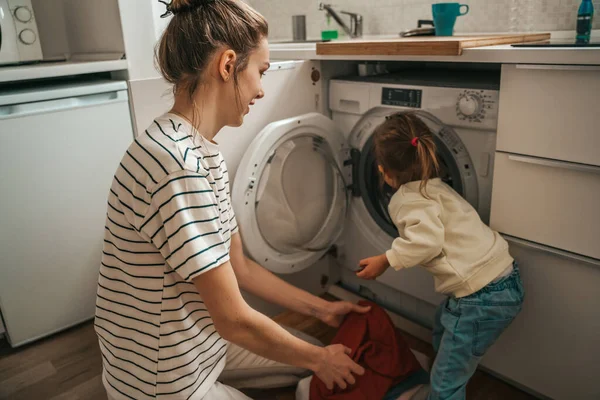Child placing the laundry items into the open washing machine assisted by her female parent