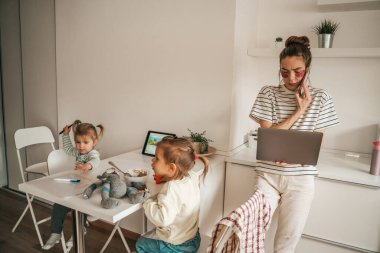 Çocukları masada oynarken laptopunu kullanan göz altı bantlı ciddi bir kadın.