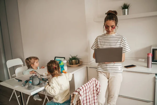 Woman with the under-eye patches working on the laptop while children watching cartoons on the tablet