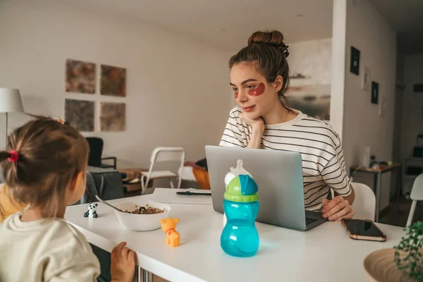 Woman with the under-eye patches seated at the kitchen table with the laptop looking at her daughter at breakfast