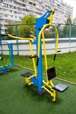 Spor sahasında parlak sarı ve mavi renkli metal bir simülatör. Yeşil ve konut binalarının arka planında lastik kaplama var. Spor sağlık hobileri.
