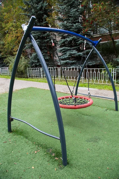 A metal swing in the form of a ring with intertwined synthetic ropes in the center against a background of greenery on a clear sunny day. Playgrounds, sports, health entertainment