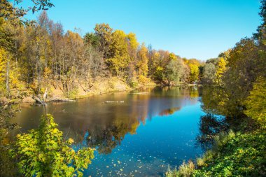 Dik kıyılarda sarı ve yeşil ağaçlarla kaplı bir gölet. Açık güneşli bir günde mavi gökyüzünün güzel bir yansıması. Doğa peyzaj nehirleri ekoloji.