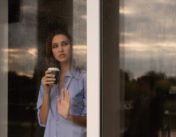 Hermosa joven con taza de café mirando a través de la ventana —  Fotos de Stock
