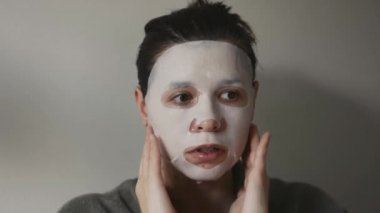 Kadın yüzünü nemlendirmek için bez maske kullanıyor. Gri arka planda izole edilmiş sağlıklı bir yaşam tarzı.