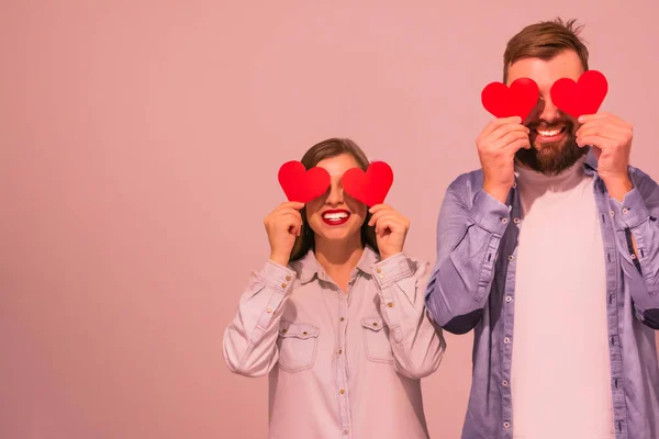 Χαρούμενο πολυφυλετικό ευτυχισμένο ζευγάρι που καλύπτει τα μάτια με κόκκινες χάρτινες καρδιές και χαμογελαστό ροζ φόντο Royalty Free Φωτογραφίες Αρχείου