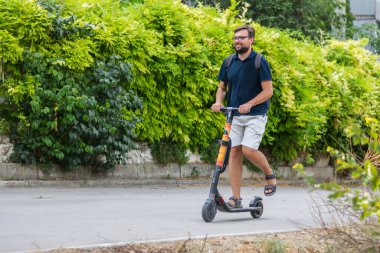 Sırt çantalı sakallı yetişkin bir adam yaz parkında ekolojik taşımada elektrikli scooter kullanıyor.